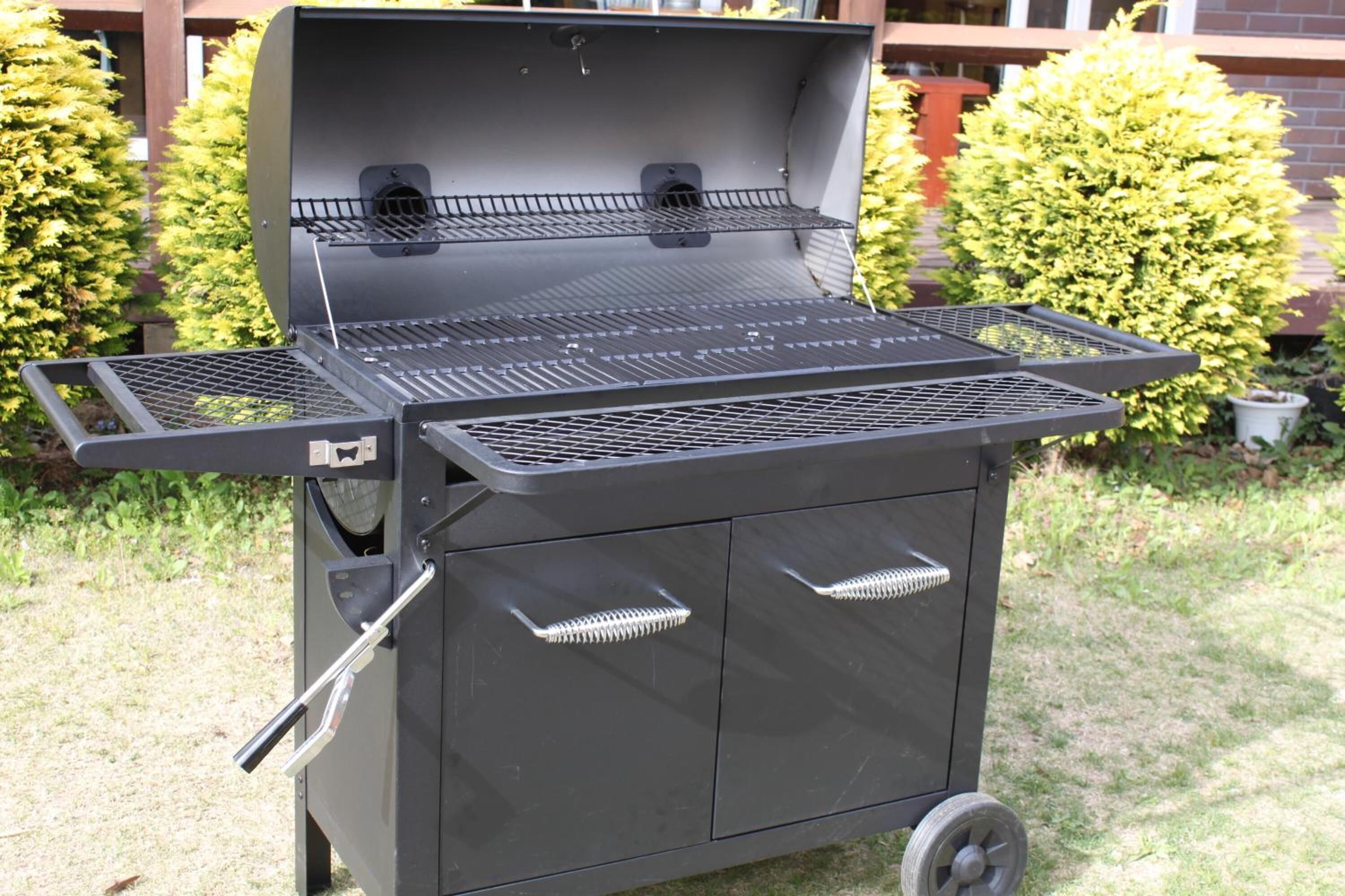 Un barbecue noir pour l’extérieur, doté d’une étagère latérale et de roulettes pour faciliter son déplacement, installé sur une pelouse avec des buissons en arrière-plan.