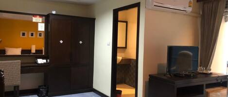 Quarto de hotel com um guarda-roupa espaçoso, uma secretária com um computador, um ar condicionado e um banheiro visível através de uma porta aberta.