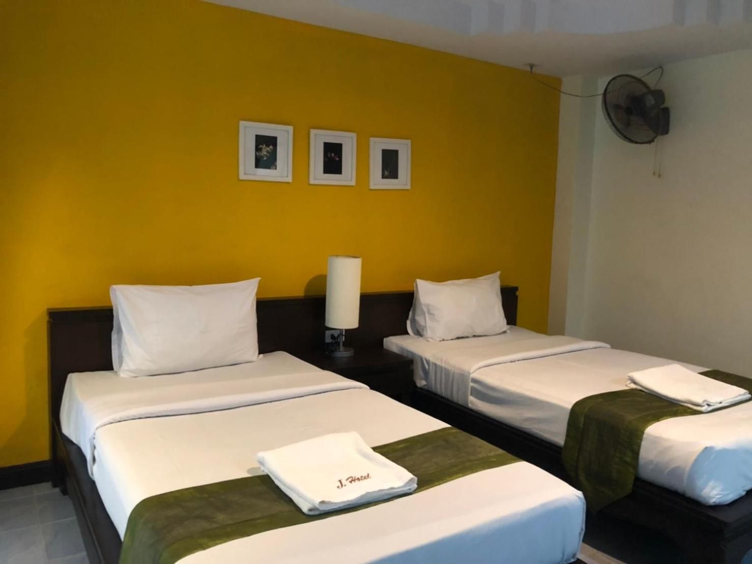 Une chambre d’hôtel avec deux lits, du linge de lit blanc, un plaid vert et un ventilateur fixé au mur.