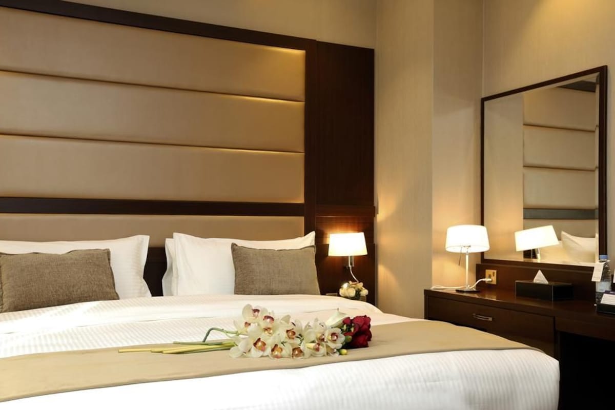 A hotel room with a large bed, two bedside lamps, a nightstand, a mirror, and a bouquet of flowers on the bed.