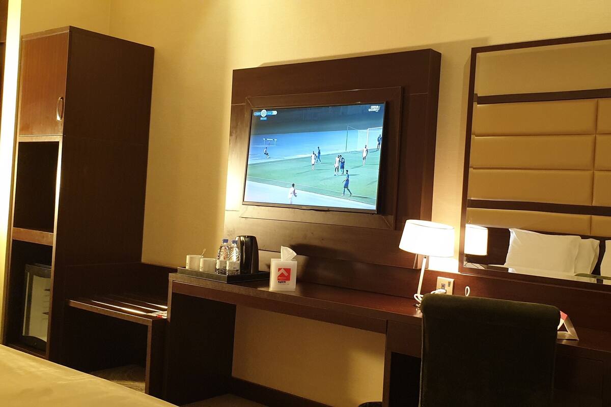 A hotel room with a TV mounted on the wall, a desk with a lamp, a chair, and a bed.