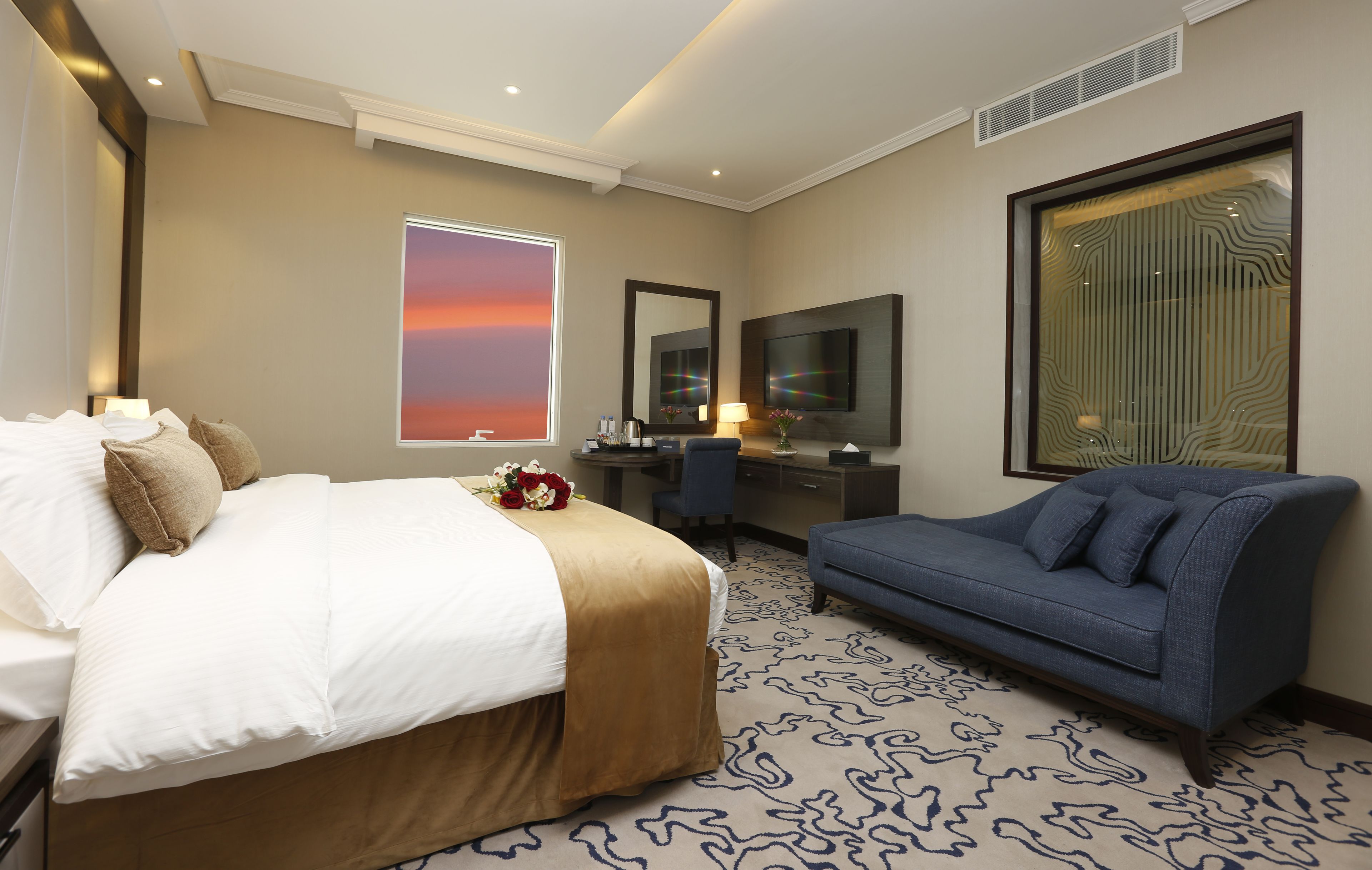 A hotel room with a bed, a desk, a sofa, a large window with a sunset view, and a TV.