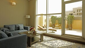 A modern living room with a grey sofa, a coffee table with a vase of flowers, and a large window with a view of a building and potted plants outside. - Swiss Flora Inn Hotel Riyadh (Riyadh)