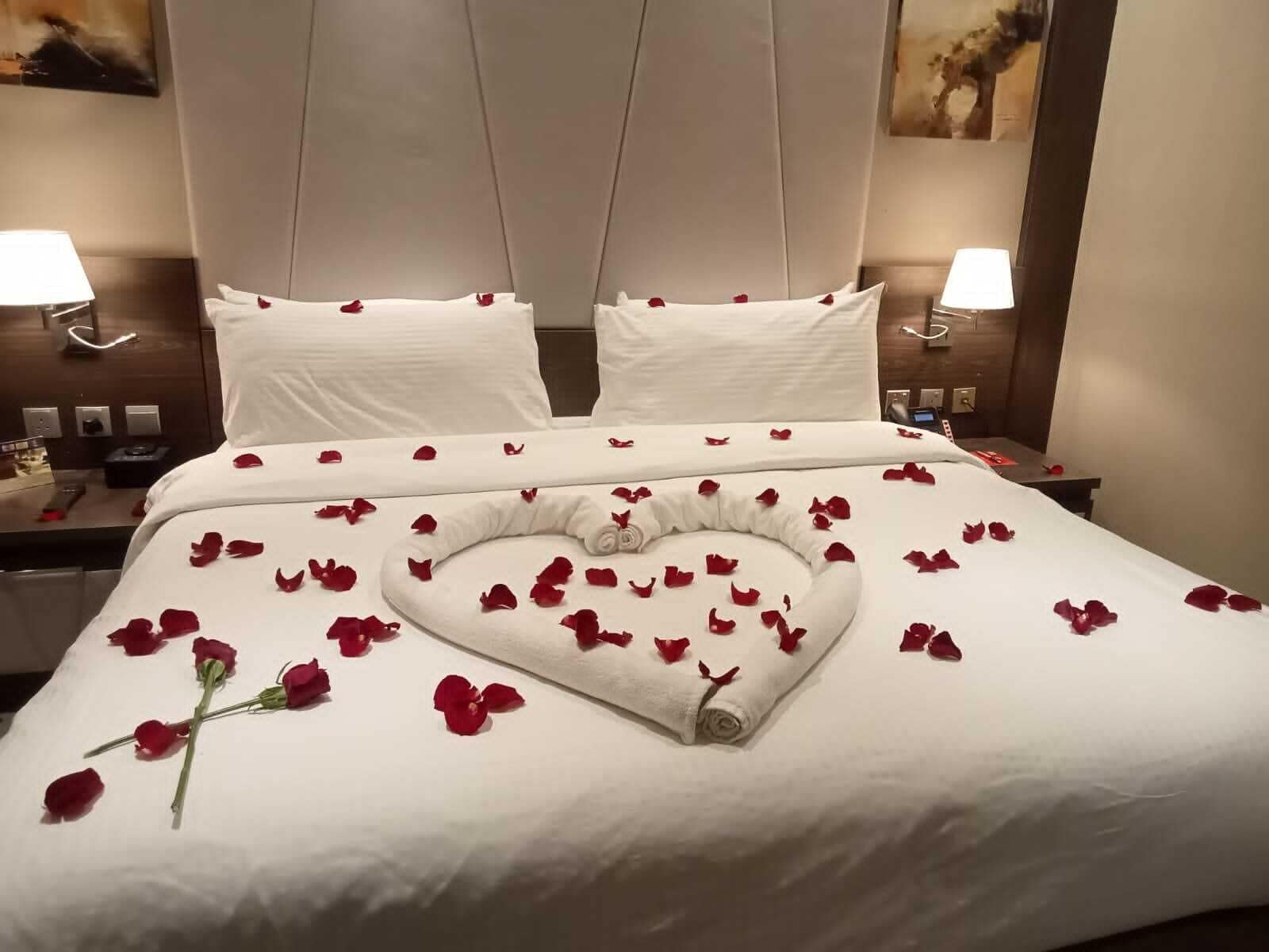 Habitació d'hotel amb un llit ben fet, roba blanca i pètals de rosa disposats en forma de cor.