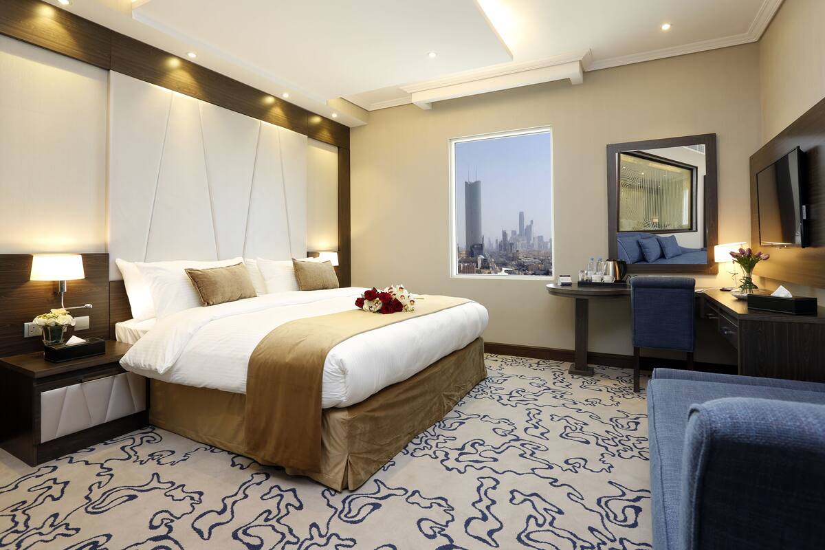 A modern hotel room with a large bed, a desk, a chair, a TV, and a window with a city view.