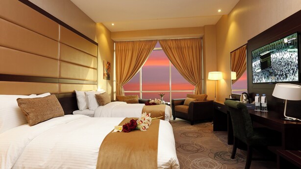 A hotel room with two beds, a desk, a chair, a TV, and a view of a sunset. - Swiss Flora Inn Hotel Riyadh (Riyadh)