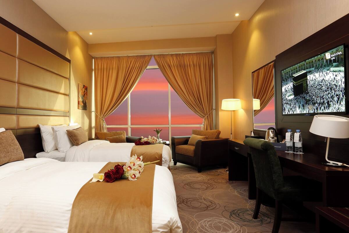 A hotel room with two beds, a desk, a chair, a TV, and a view of a sunset.