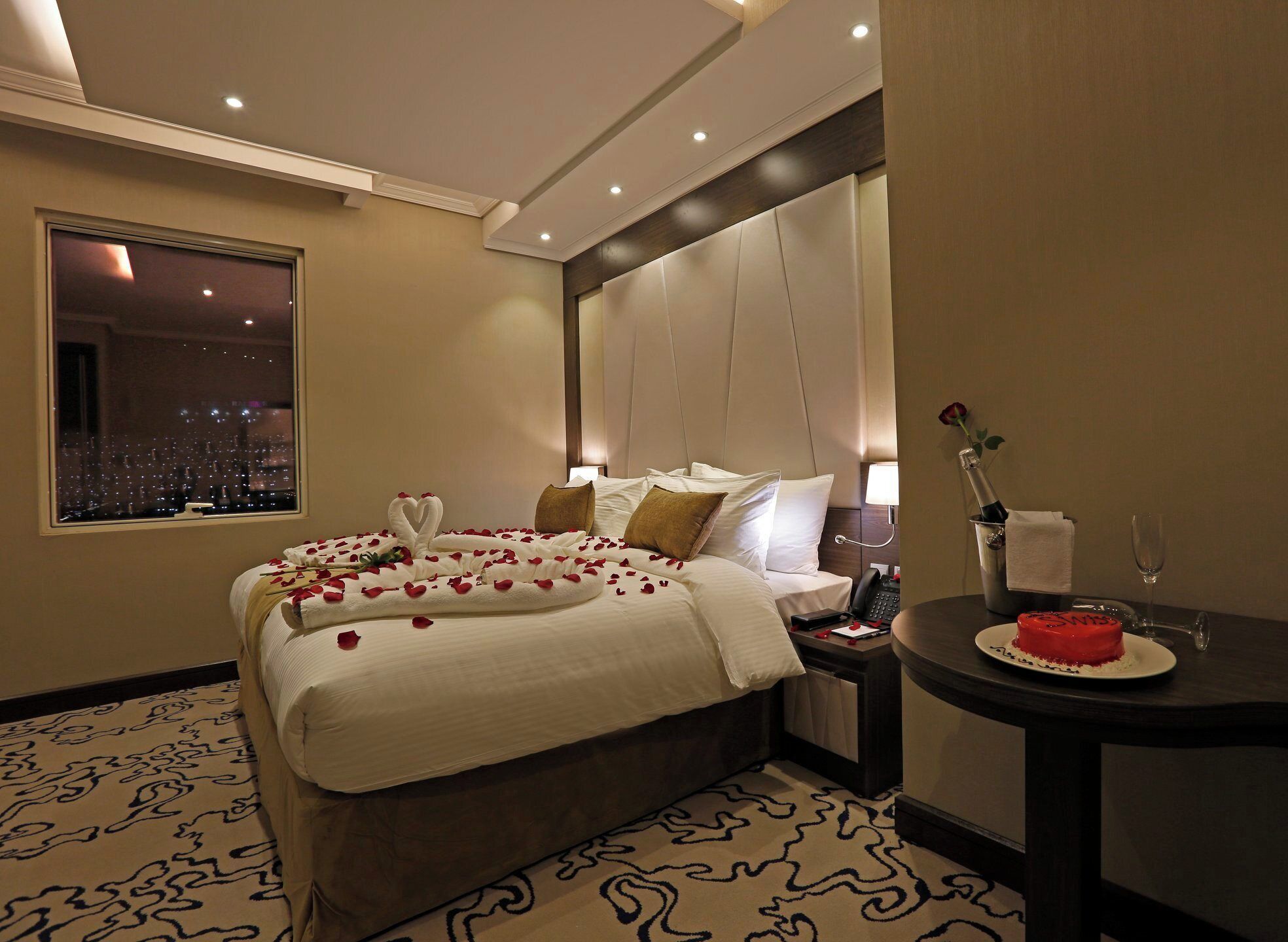 A hotel room with a large bed, a nightstand, a small table with a red cake, and a view of a cityscape through a window.