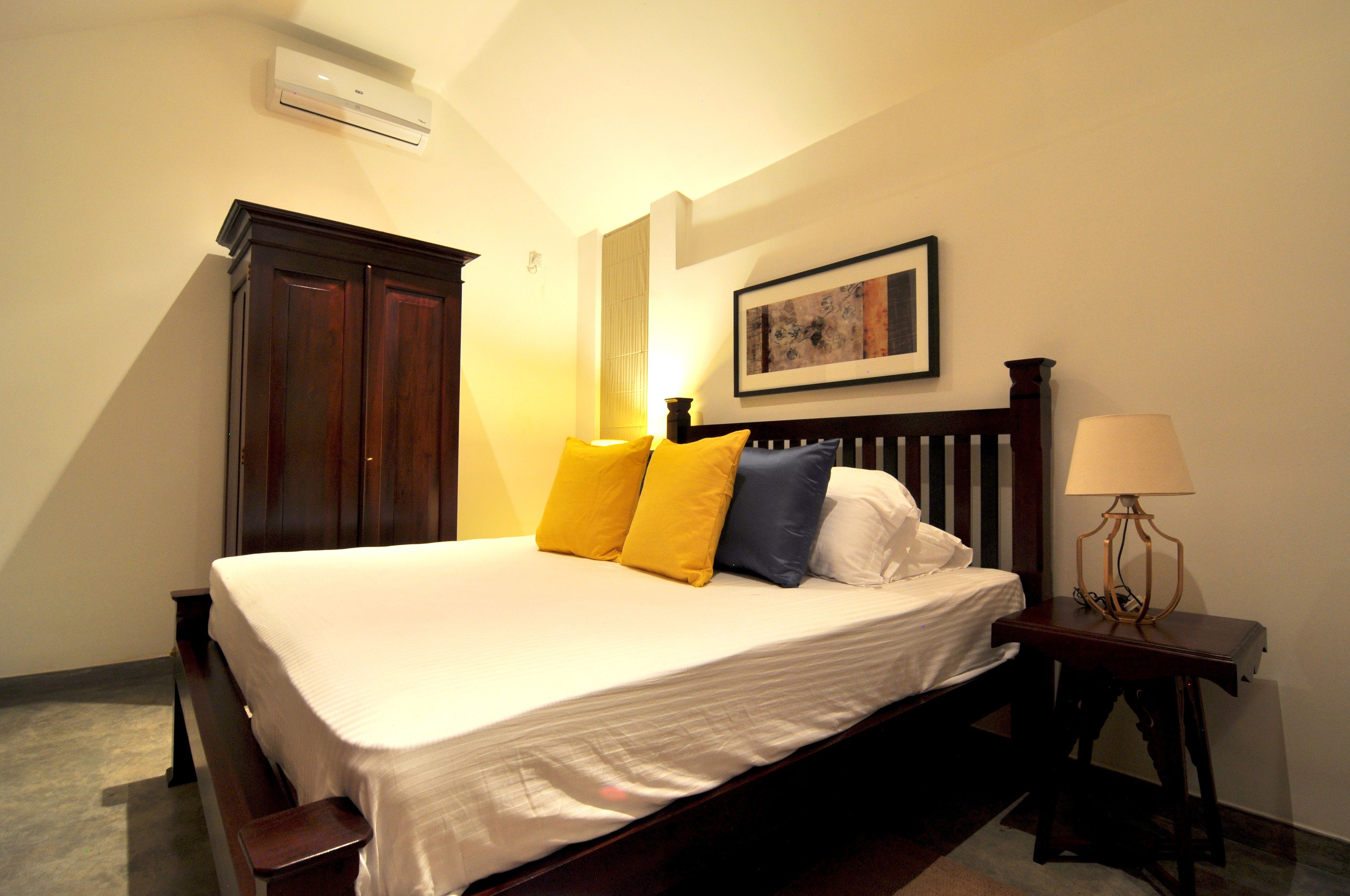 A neatly made bed with a white comforter and three pillows in yellow, blue, and white. A wooden nightstand with a lamp is beside the bed.