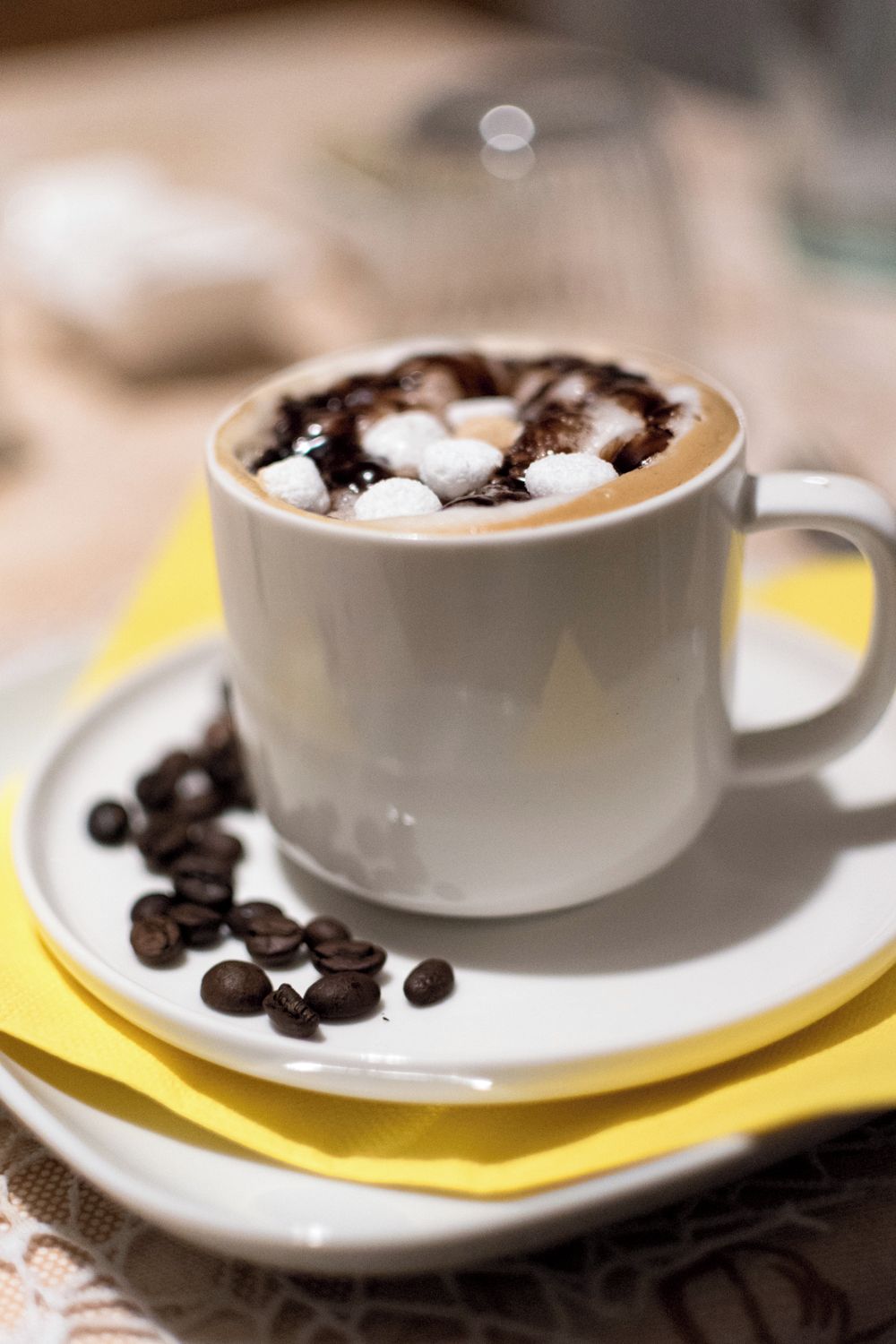 Eine weiße Tasse heiße Schokolade mit Marshmallows auf einem Untertaler, umgeben von verstreuten Schokoladenbohnen auf einer gelben Serviette.