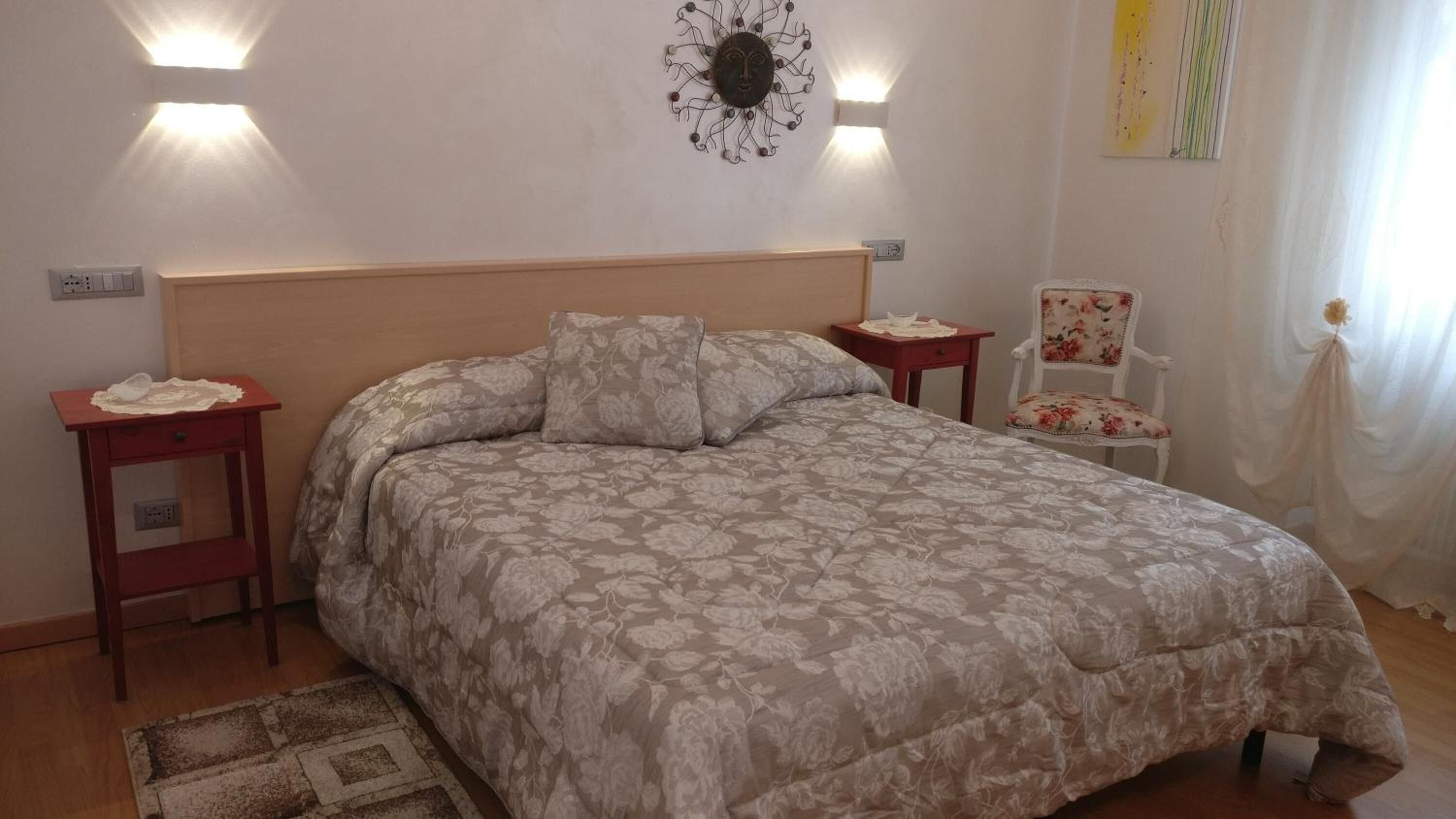 A bedroom with a floral-patterned bedspread, a wooden headboard, two bedside tables, a nightstand, a chair, and a wall-mounted light fixture.