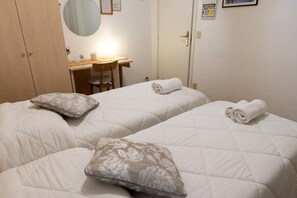 Una habitación de hotel con dos camas, cada una con una almohada y una toalla enrollada. Hay un escritorio con una silla y un espejo encima.