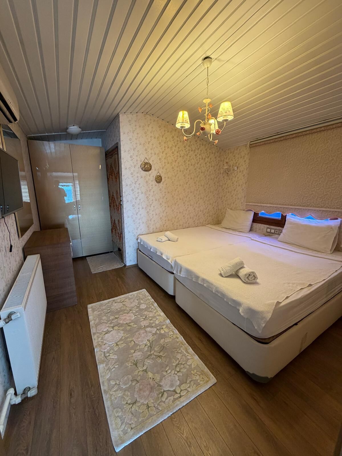 Een slaapkamer met een bed, nachtkastje, radiator, een vloerkleed met bloemenprint en een plafond met een lamp.