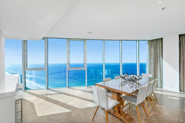 A modern dining area with a large table and chairs, large windows with ocean views, and a high ceiling.