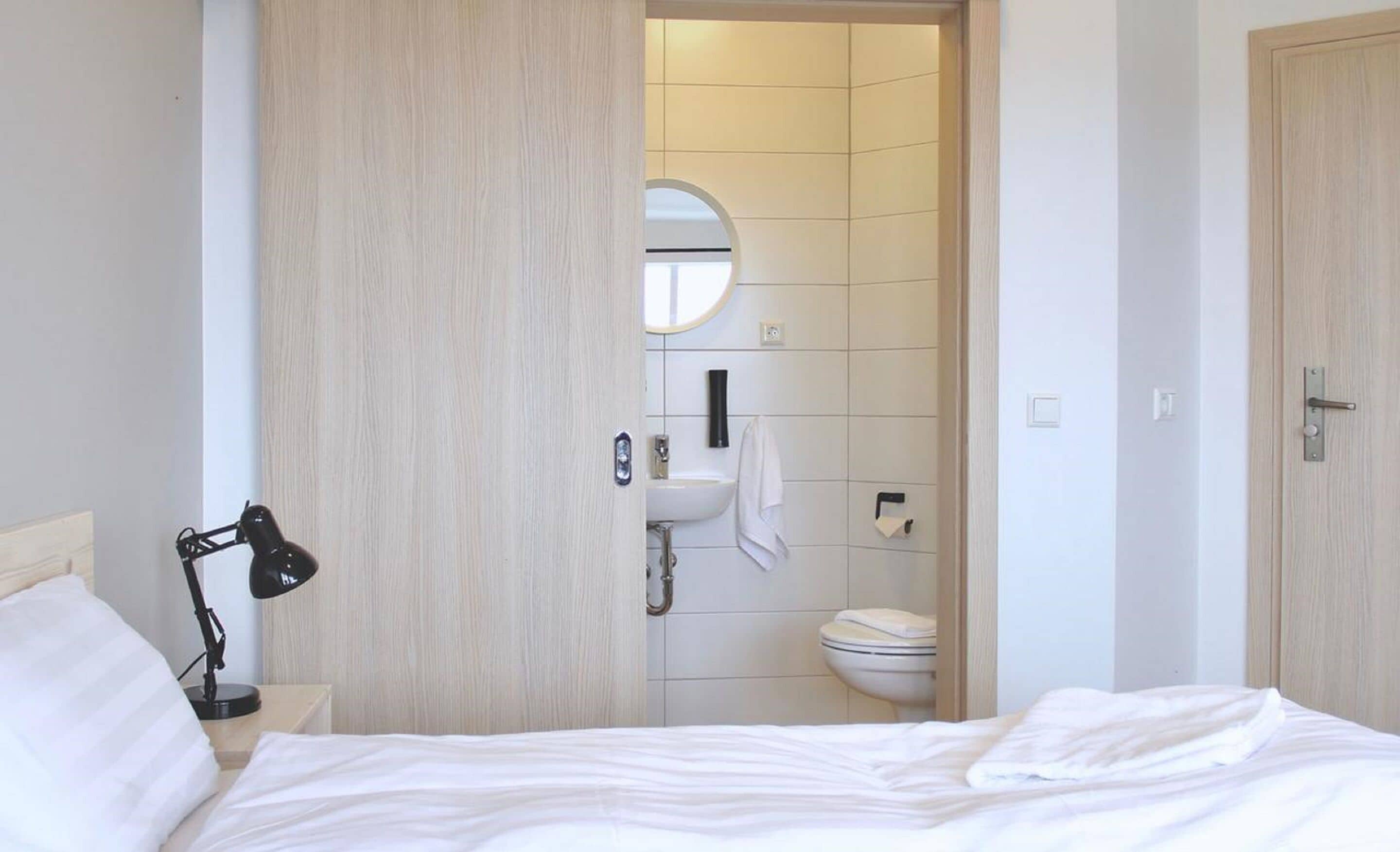 Une chambre à coucher avec un lit, une lampe de chevet, une armoire en bois et une salle de bain visible à travers une porte ouverte.