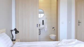 Une chambre à coucher avec un lit, une lampe de chevet, une armoire en bois et une salle de bain visible à travers une porte ouverte.