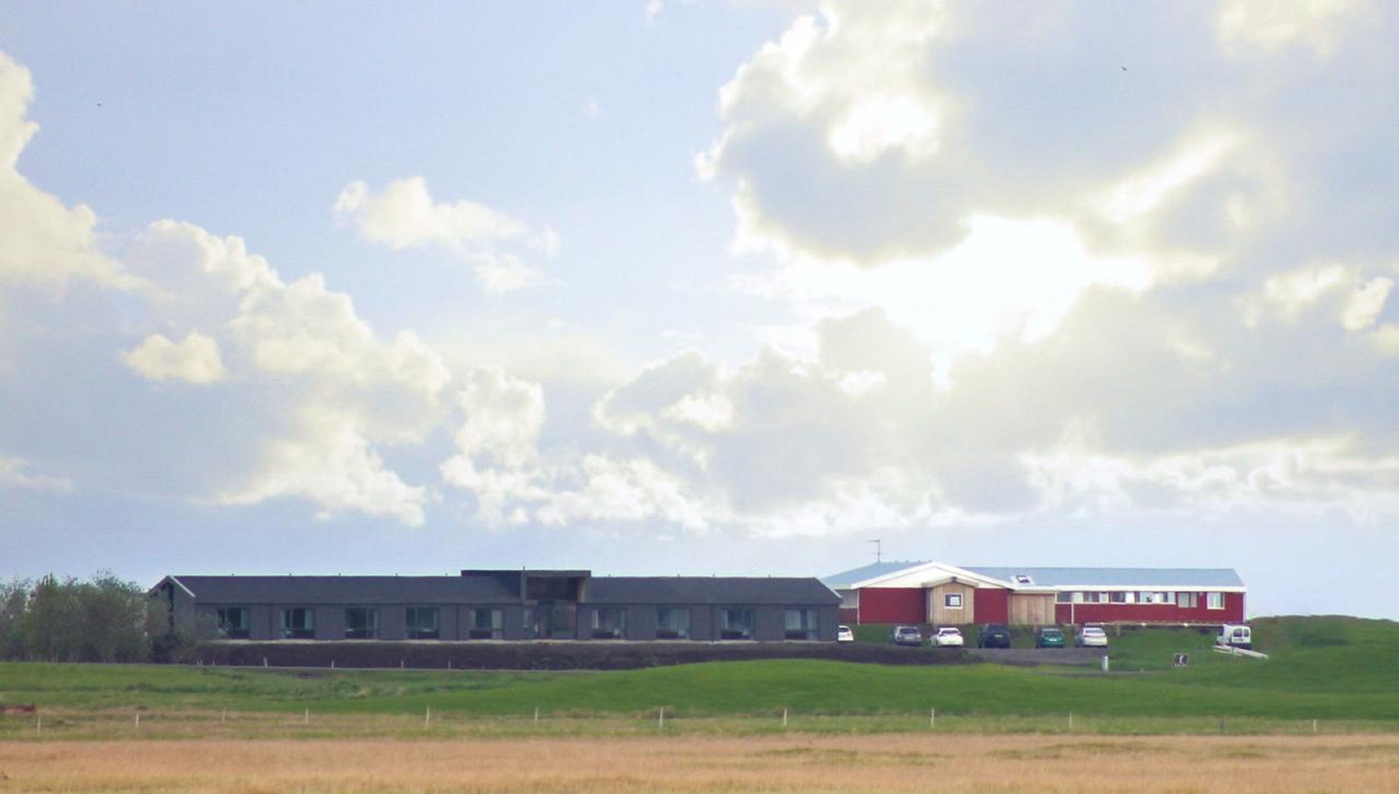 A large building with a flat roof, a red section on the right, and a parking area with several cars.