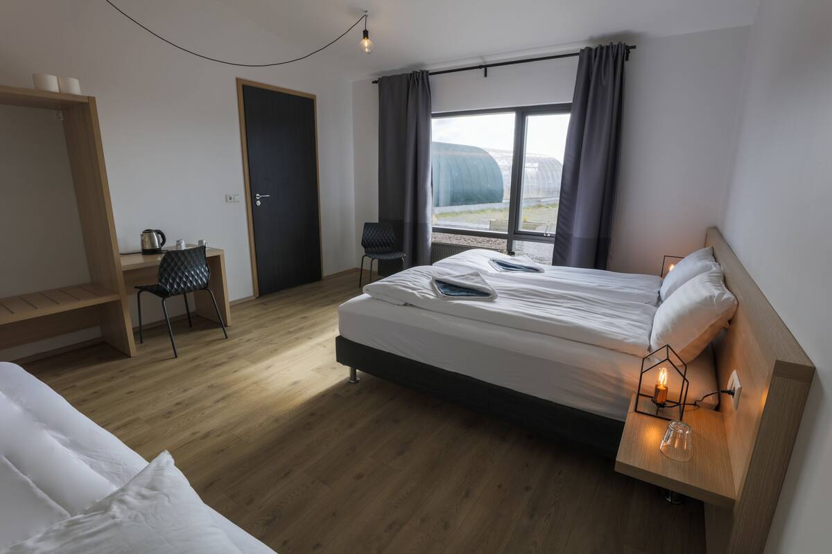 A modern hotel room with a wooden floor, a large bed with white linens, a desk with a chair, and a view of a greenhouse through the window.