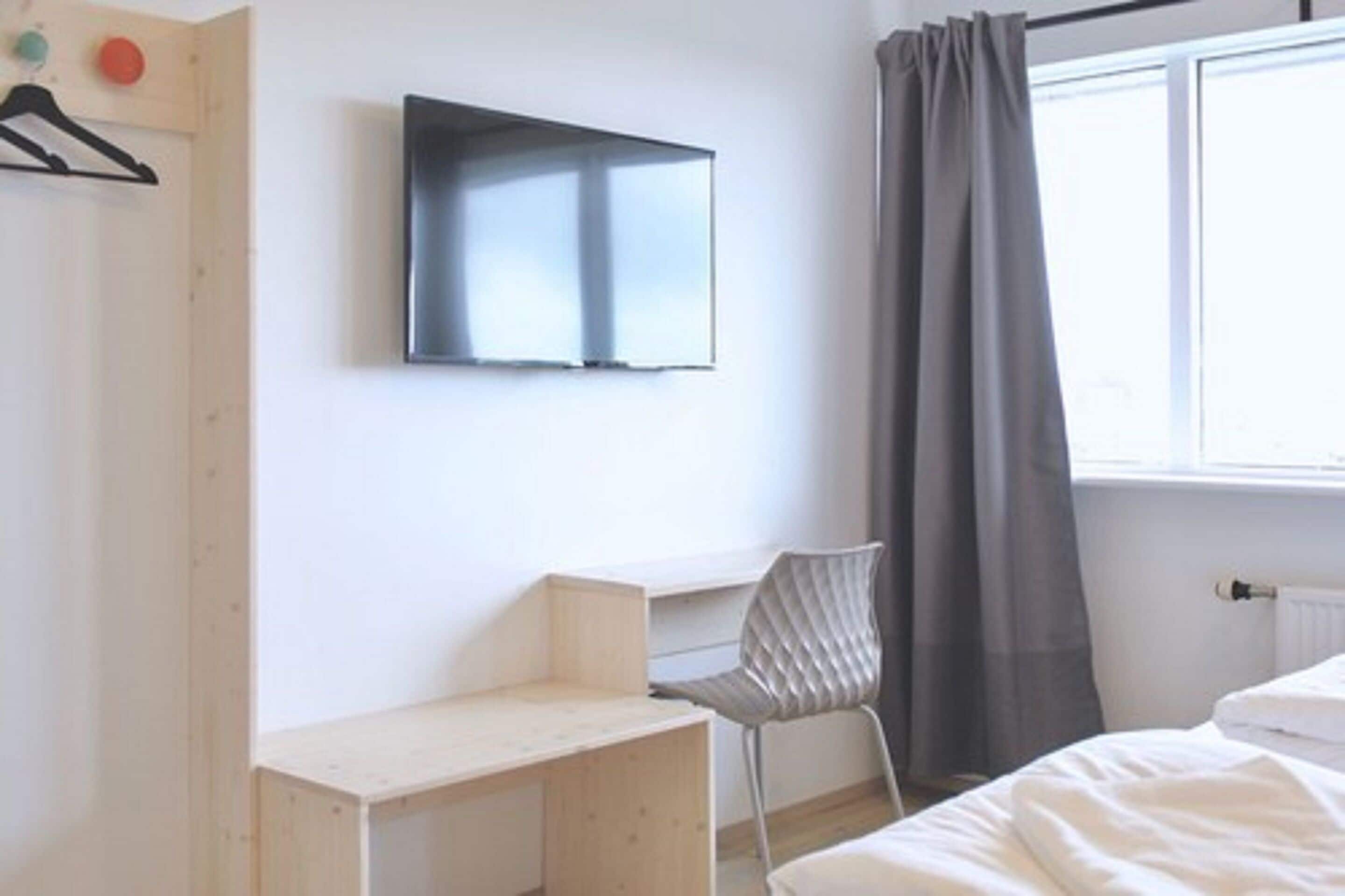 A minimalist bedroom with a bed, a desk, a chair, a TV, and a window with curtains.