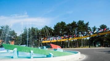 A fountain with water jets, surrounded by a green mosaic structure, and a row of flags on a yellow bridge.