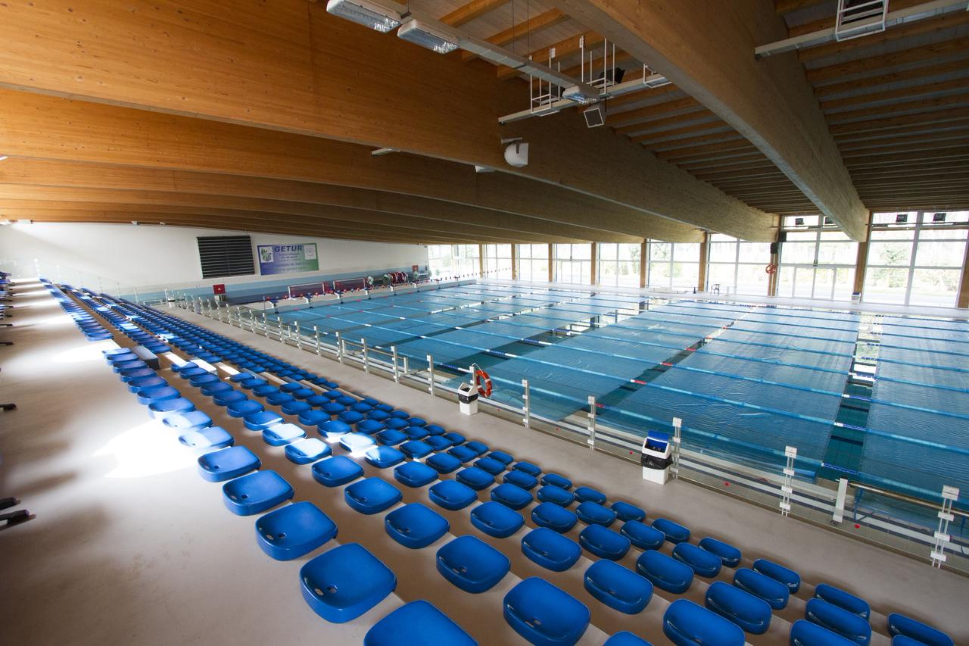 Una amplia piscina cubierta con múltiples carriles, asientos azules a los lados y techo de madera.