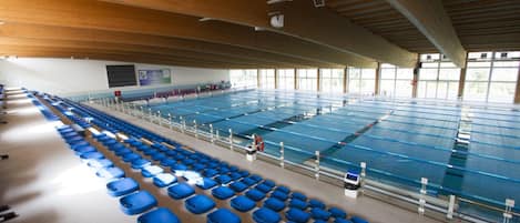 Una amplia piscina cubierta con múltiples carriles, asientos azules a los lados y techo de madera.