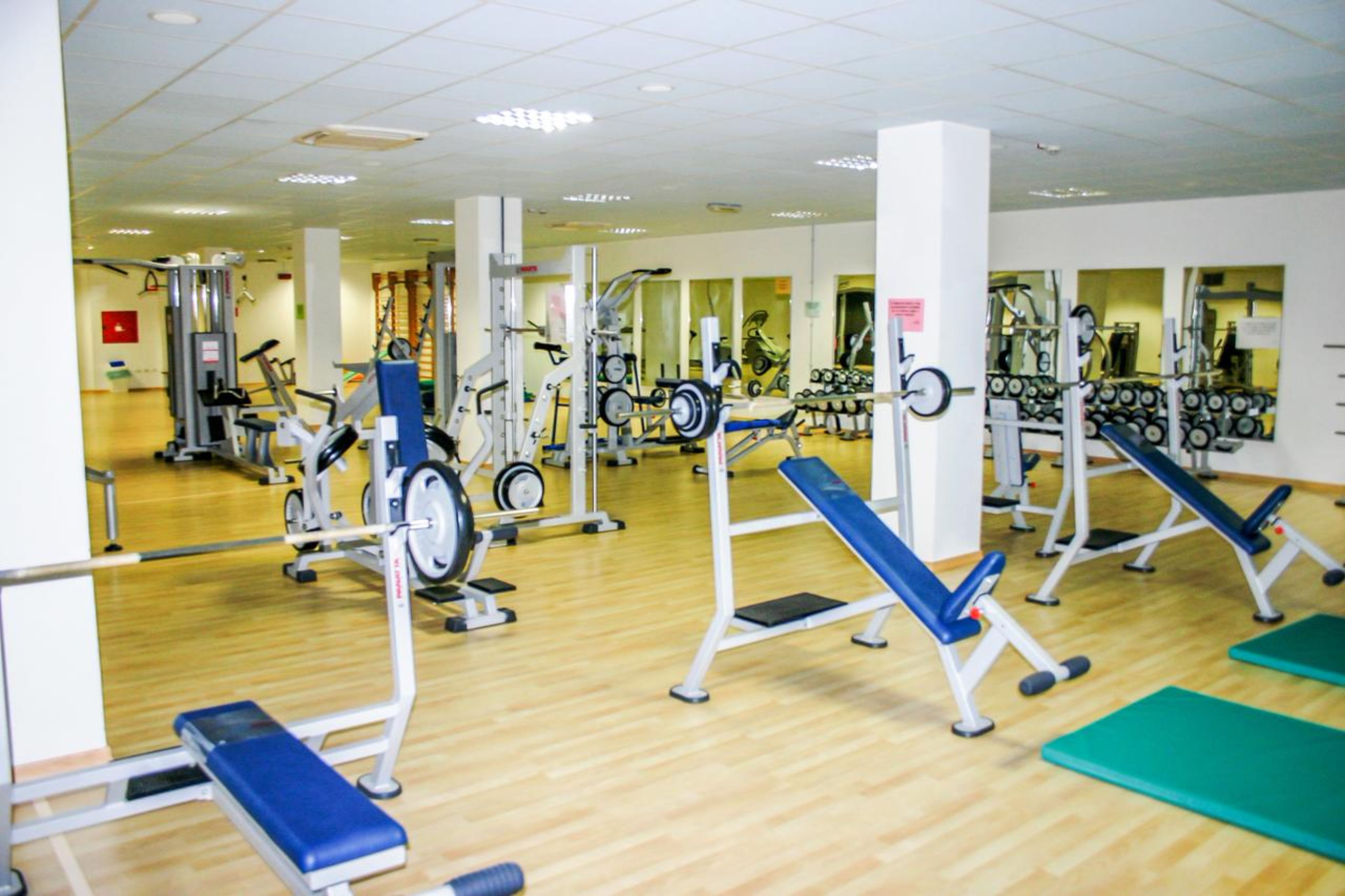 Ein Fitnessstudio mit verschiedenen Trainingsgeräten, Holzboden und Spiegeln an den Wänden.
