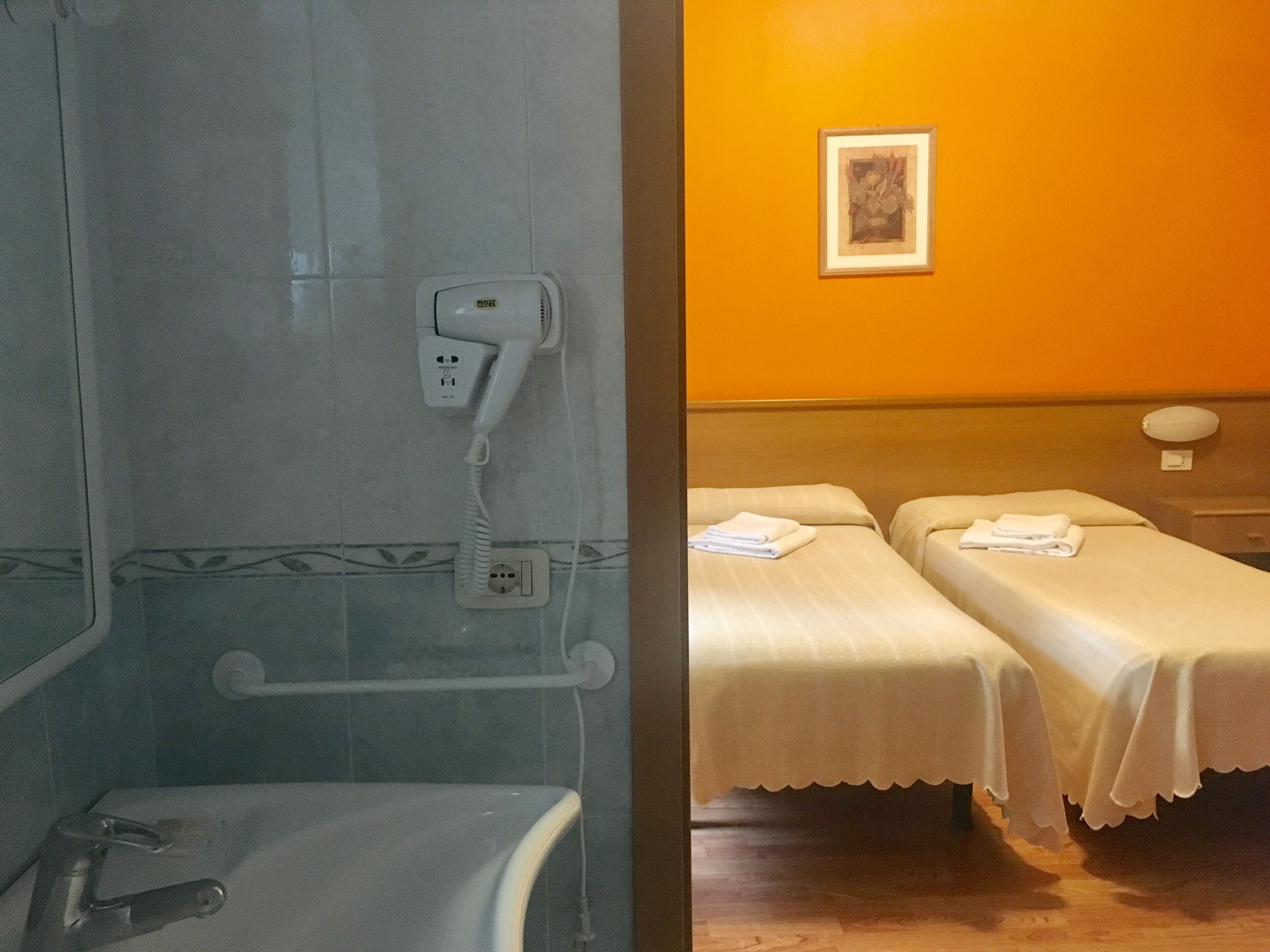 Ein Badezimmer mit Dusche, Toilette und einem Föhn, der an einer Wandsteckdose befestigt ist. Ein Bett mit weißer Bettdecke und ein gerahmtes Bild an der Wand sind ebenfalls vorhanden.
