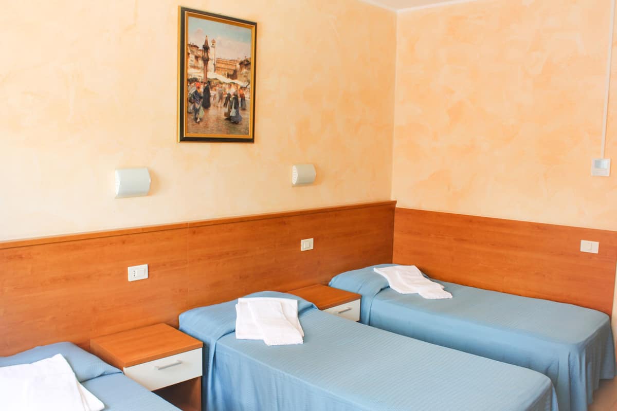 A hotel room with two single beds, wooden headboards, a small bedside table with a lamp, and a painting on the wall.