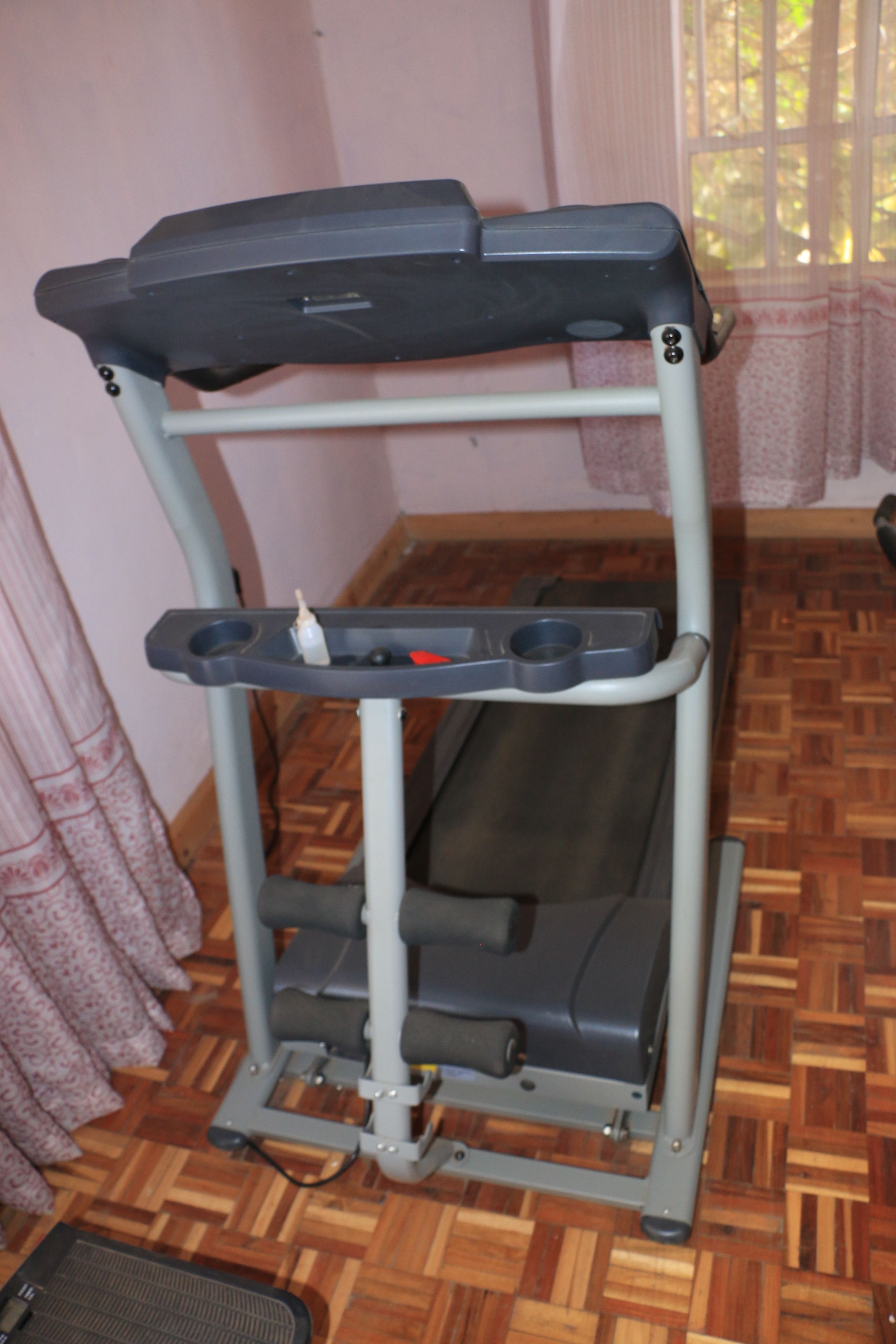 A stationary exercise bike with a tray table and foot pedals, positioned in a room with wooden flooring and a window with curtains.