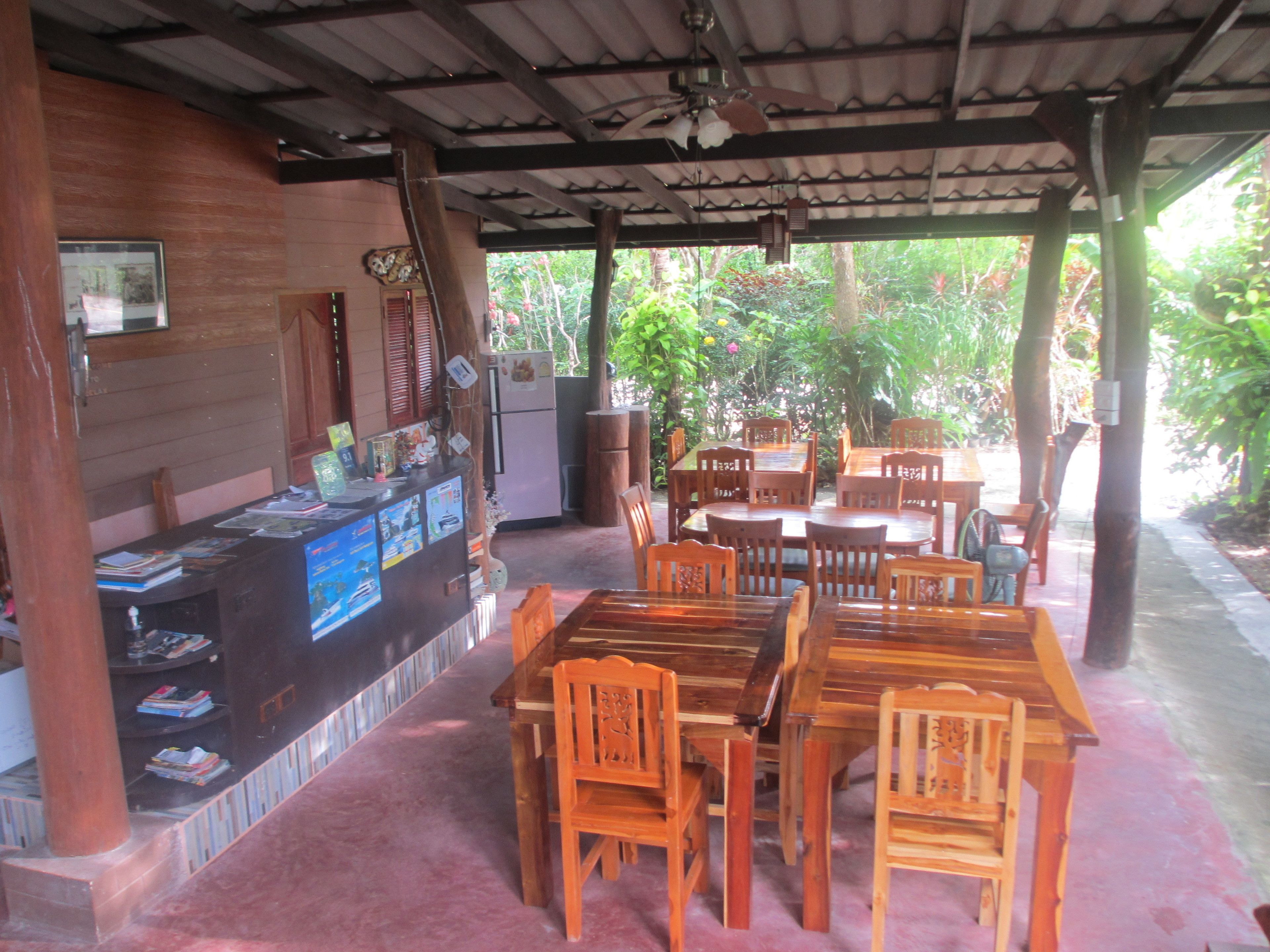 A covered outdoor dining area with wooden tables and chairs, a refrigerator, and a counter with various items.