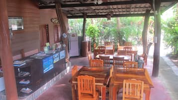 A covered outdoor dining area with wooden tables and chairs, a refrigerator, and a counter with various items.