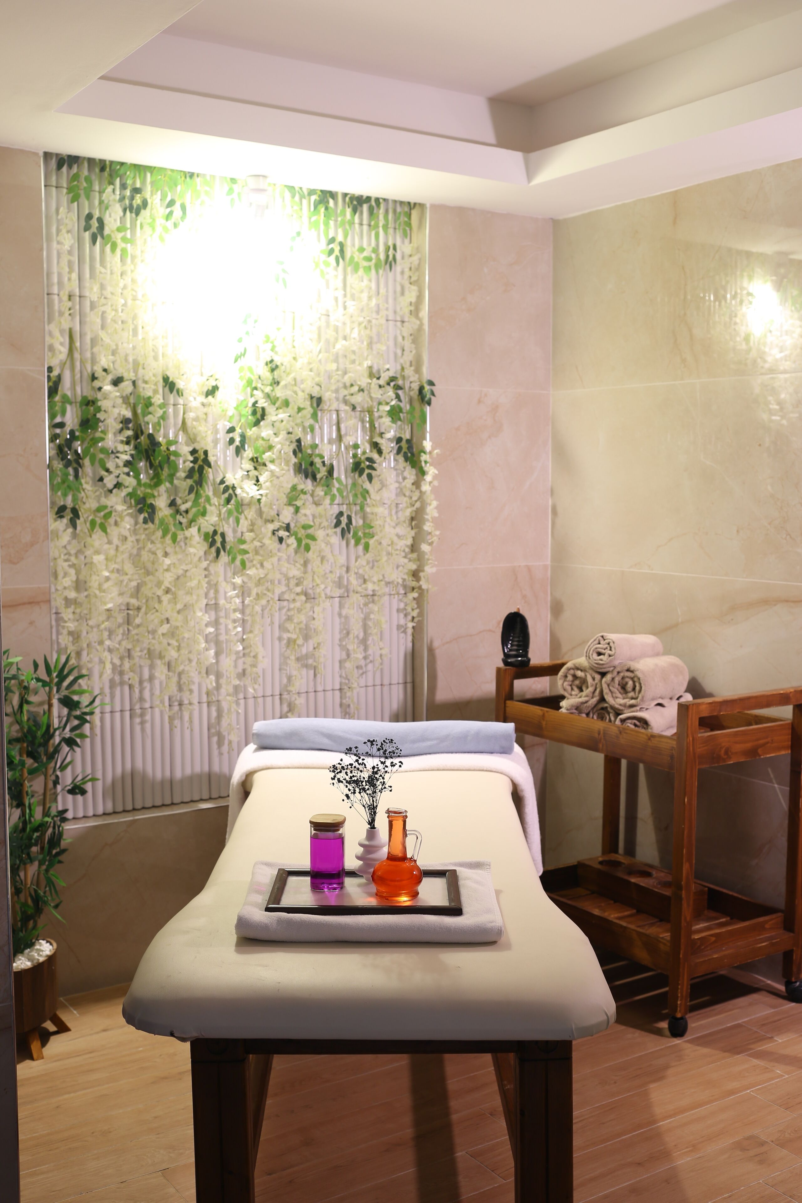 Une salle de spa équipée d’une table de massage, d’un banc en bois, d’une étagère avec des serviettes et d’un mur orné de plantes suspendues.