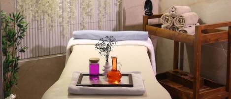 Un cuarto de spa con una camilla de masaje, un banco de madera, un estante con toallas y una pared con plantas colgantes.