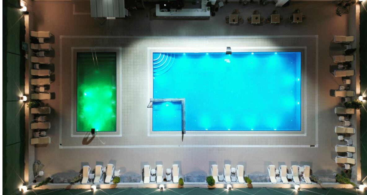 Una piscina con dos áreas diferenciadas, una con luz verde y la otra con luz azul. Hay sillas de descanso y una pequeña zona de estar con plantas.