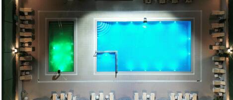 Una piscina con dos áreas diferenciadas, una con luz verde y la otra con luz azul. Hay sillas de descanso y una pequeña zona de estar con plantas.