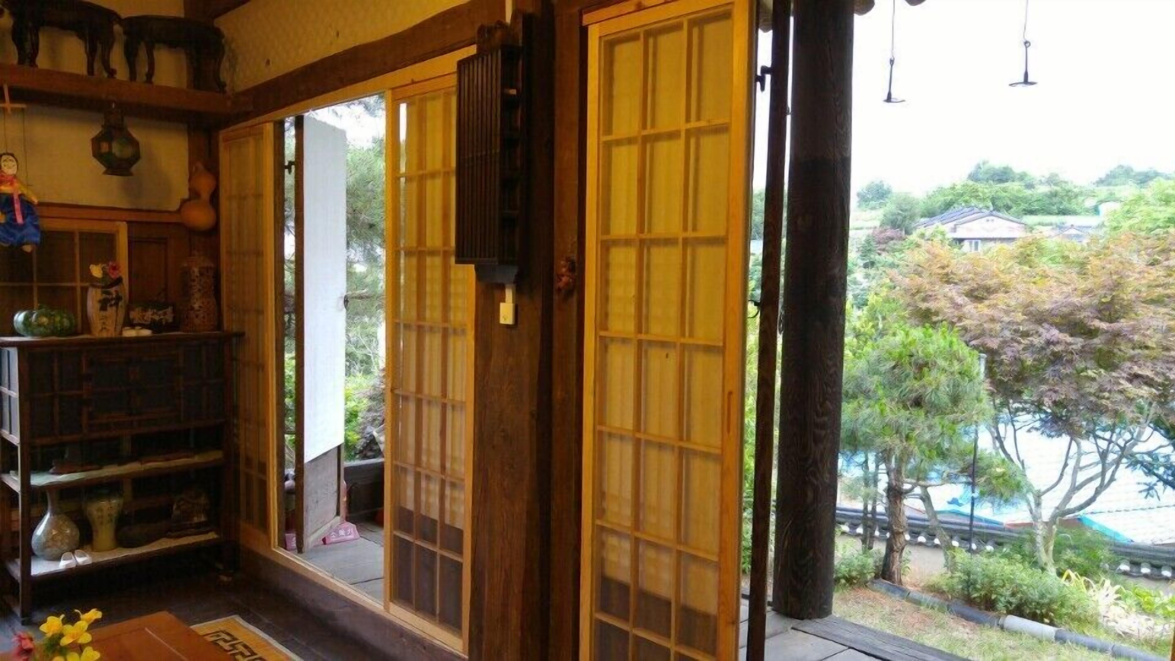 Une pièce de style japonais traditionnel, avec des portes coulissantes, une étagère en bois où sont disposés divers objets, et une vue sur un jardin.