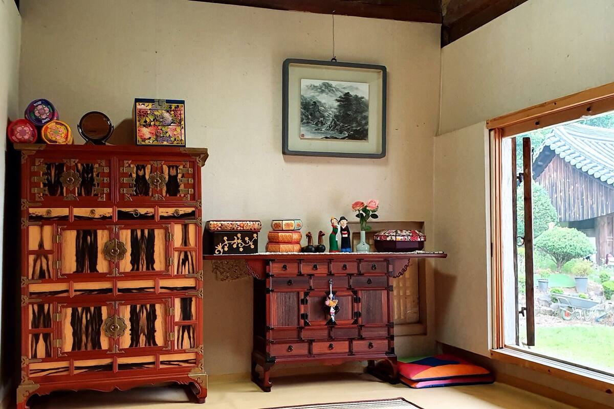 A traditional Japanese room with a wooden cabinet, a small table with decorative items, and a window with a view of a garden.