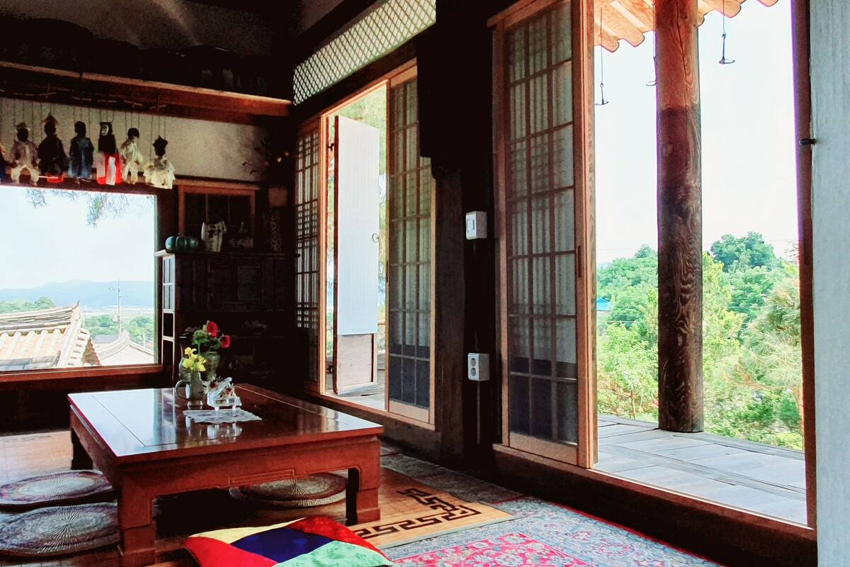A traditional Japanese room with tatami mats, a low wooden table, and sliding doors leading to a balcony with a view of a traditional building and greenery.