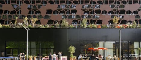 A modern building with a unique geometric facade, outdoor seating area, and a café with patrons.