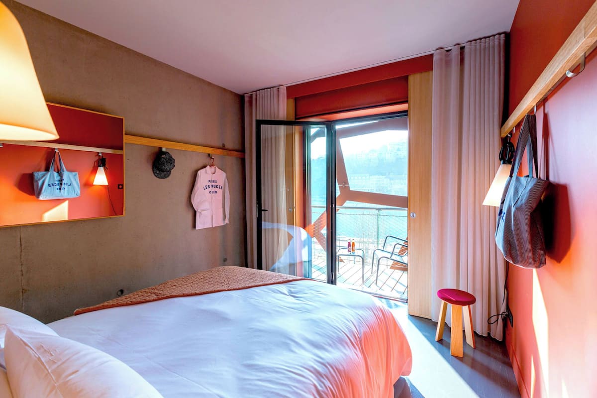 Une chambre d’hôtel avec un lit, un petit tabouret, un sac suspendu et une vue sur l’extérieur.