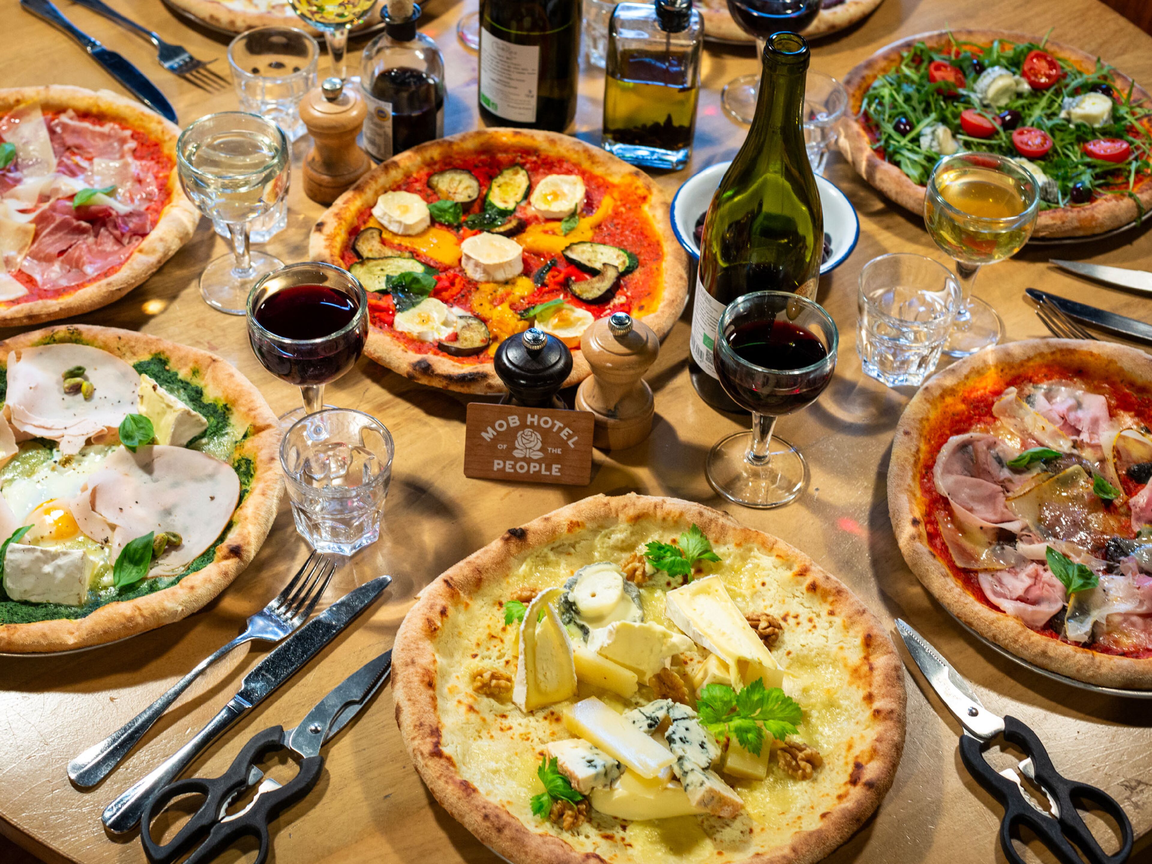 Una mesa con varias pizzas, copas de vino y cubiertos.