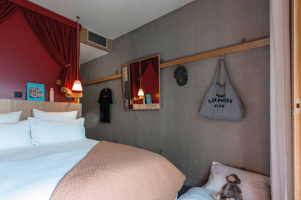 Une chambre d’hôtel avec un grand lit, un plus petit lit, un miroir, un sac suspendu et un jouet en peluche.