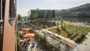 Un edificio moderno con fachada de vidrio que da a la ribera, con mesas al aire libre y zonas verdes.