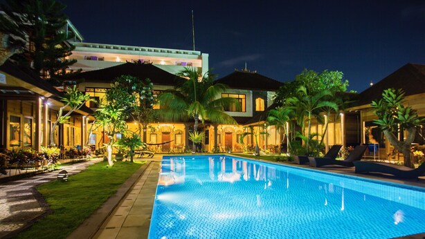 A swimming pool with a tiled deck, surrounded by palm trees and a building with multiple balconies. - Easy Surf Camp (Kuta)