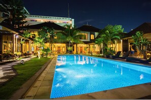 A swimming pool with a tiled deck, surrounded by palm trees and a building with multiple balconies. - Easy Surf Camp (Kuta)