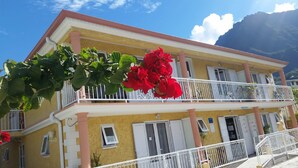 Un bâtiment jaune doté d’un balcon, de volets blancs et de fleurs rouges.