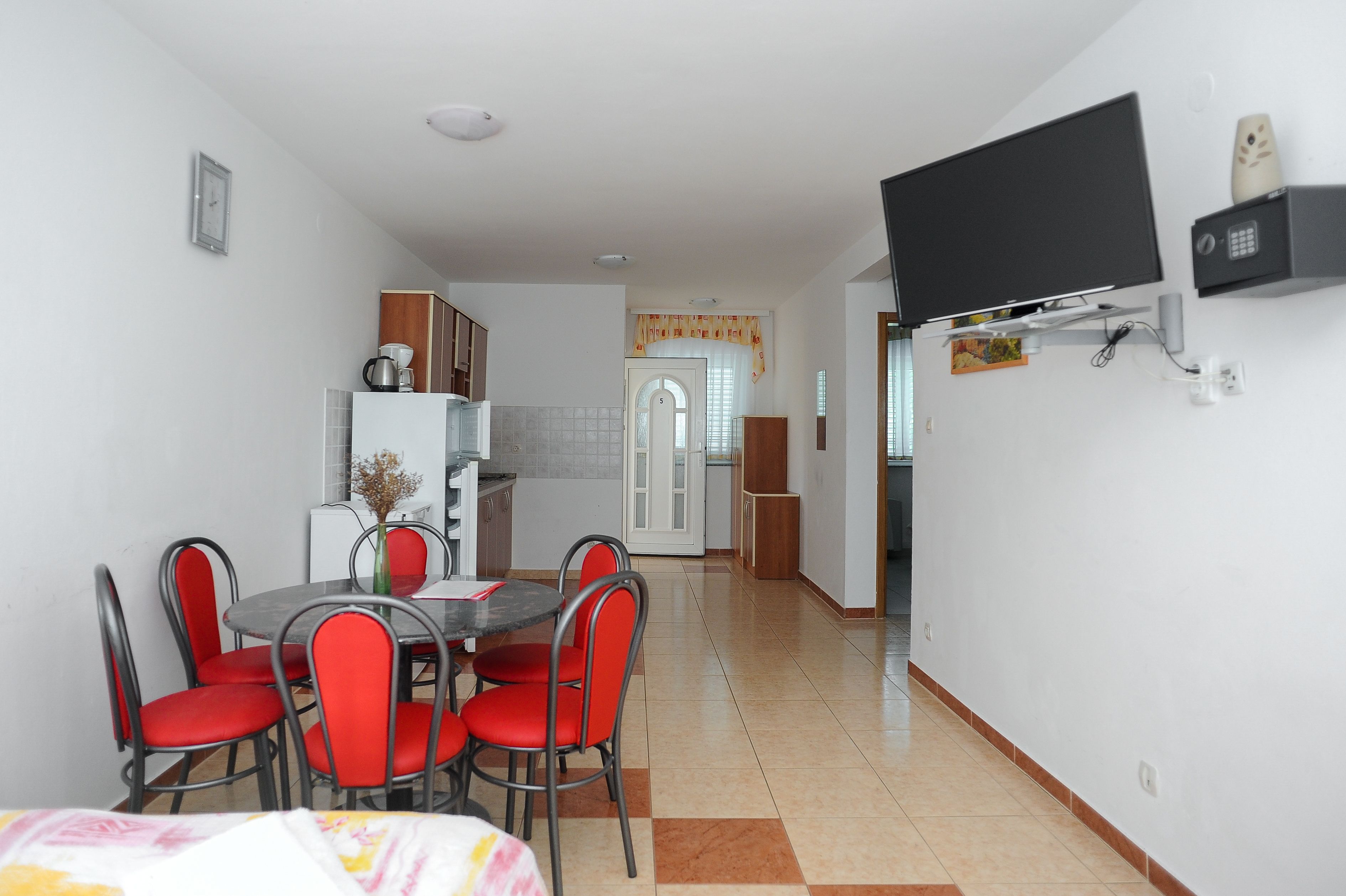 A compact living space with a dining area featuring a round table and red chairs, a kitchen with a refrigerator and cabinets, and a mounted television on the wall.