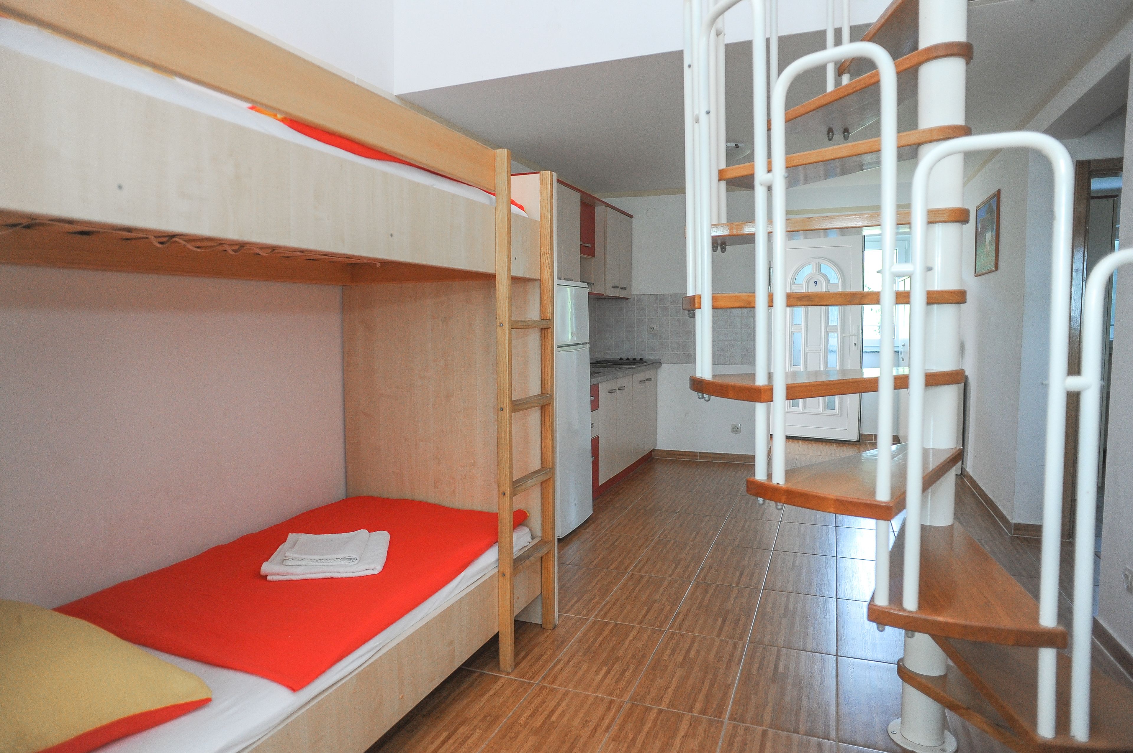 A bunk bed with a red mattress, a wooden ladder, and a kitchen area with cabinets and a refrigerator.
