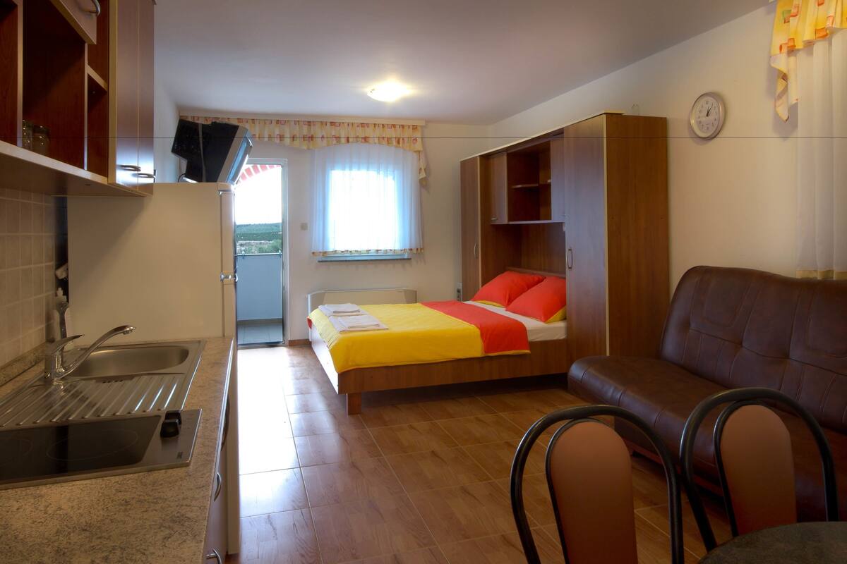 Ein gemütlicher Raum mit Küchenzeile, einem Bett mit roten Kissen, einem braunen Ledersofa und einem Fenster mit Vorhängen.